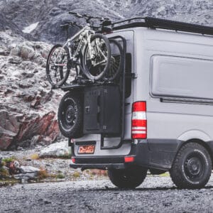 The rear part of a W906 fully grey raptored Sprinter van. The rear of the van is equipped with black powdercoated, aluminium exterior gear. Such as a Tire Carrier, Cargo Ladder, Cargo Box and a Cargo Frame carrying two mountainbikes. In the backgroudn you see rocky Swiss mountain terrain.
