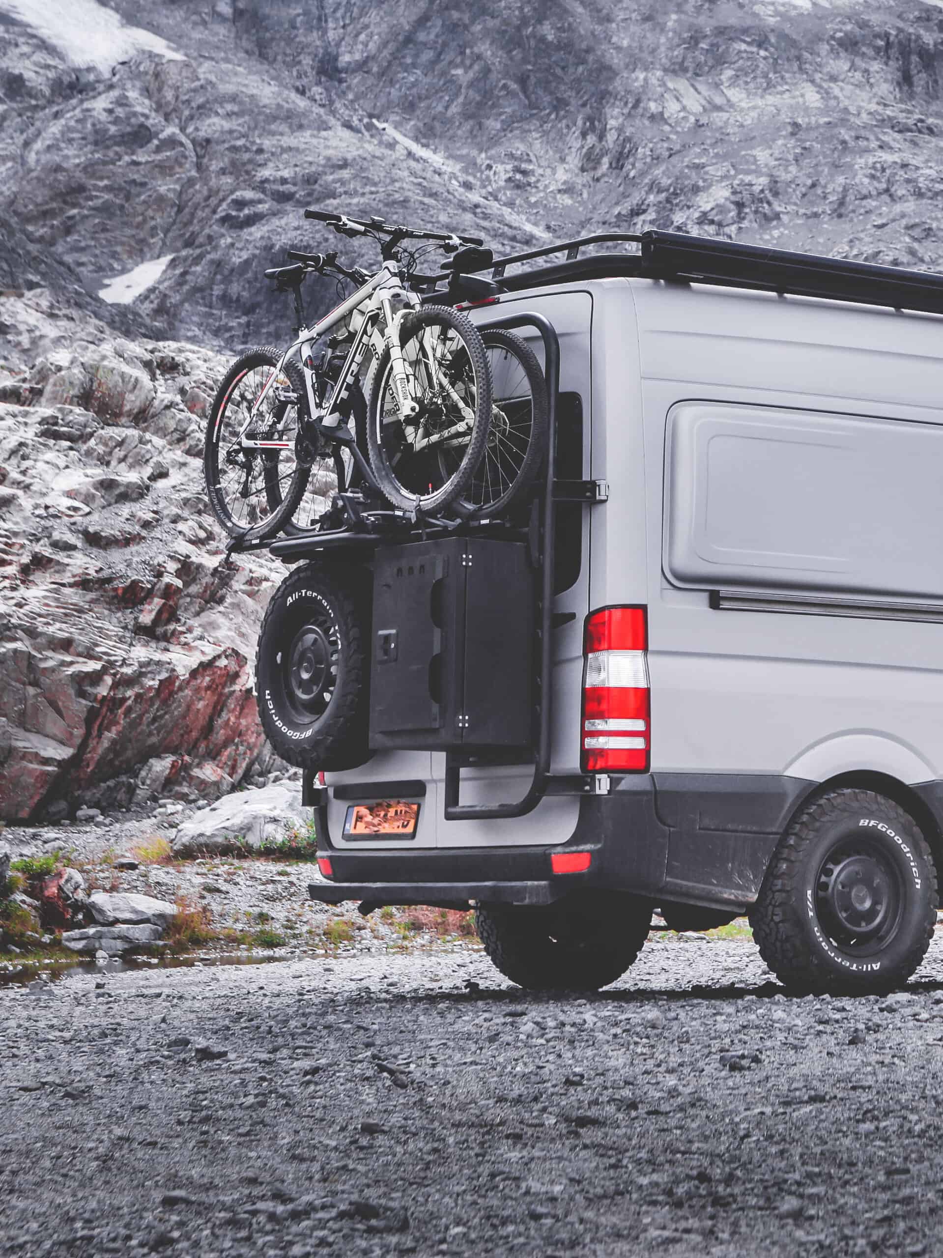The rear part of a W906 fully grey raptored Sprinter van. The rear of the van is equipped with black powdercoated, aluminium exterior gear. Such as a Tire Carrier, Cargo Ladder, Cargo Box and a Cargo Frame carrying two mountainbikes. In the backgroudn you see rocky Swiss mountain terrain.