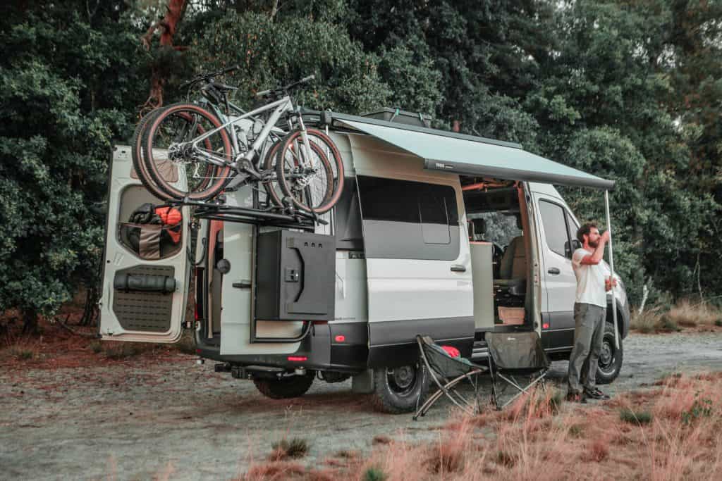 The pebble grey Mercedes Sprinter 4x4 is parked in a forrest. The owner is roling out the awning. The side and rear doors are open and two camping chairs are position underneath the awning. The campervan is equipped with all Dutchvanparts exterior and SOLO interior.