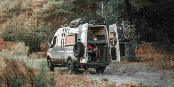 Pebble grey Mercedes Sprinter 4x4 rear side view in the forest. The rear doors are open and inside you see a lot of outdoor and sports equipment such as kits, helmets and yoga mats. The outside of the van is equipped with exterior gear.