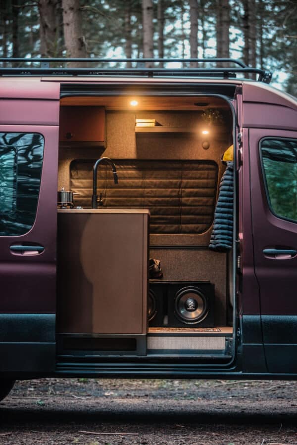 The side view of a burgundy 4x4 Mercedes Sprinter campervan with the sliding door open. Looking into the van you see a dark brown aluminium interior with cozy lights on.